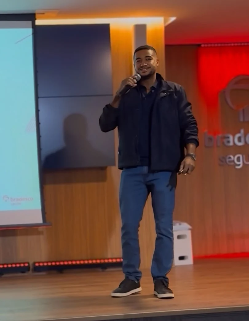Jonatan Jamar no palco do auditório da Bradesco Seguros, de microfone na mão, apresentando para o time de Operações Saúde